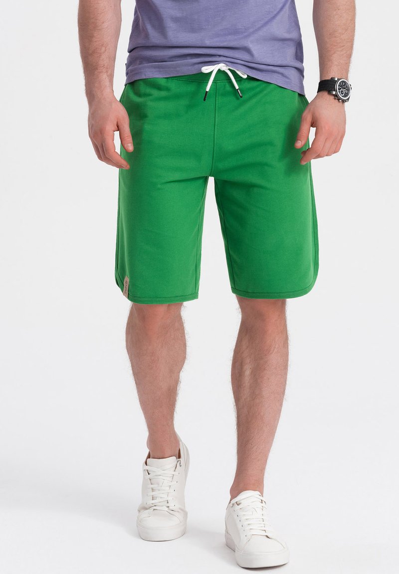 Shorts en coton vert avec une taille élastique et un cordon blanc, présentant une coupe droite et une petite étiquette de marque sur le côté.
