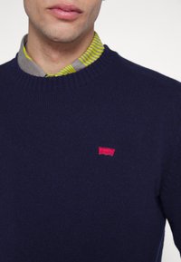 Maglione girocollo blu realizzato in materiale tessuto, con un piccolo patch logo rosso. È visibile un colletto multicolore a righe sotto.