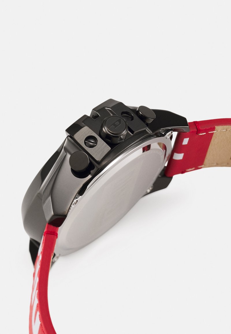 Fekete fém karóra tok, két kerek gombbal és egy koronával, piros bőr pántba rögzítve, fehér varrással.