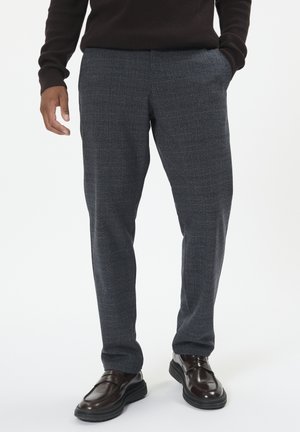 Dunkelgraue Karottenhose aus strukturiertem Stoff, mit lässiger Passform und Seitentaschen, kombiniert mit glänzenden schwarzen Slippern.