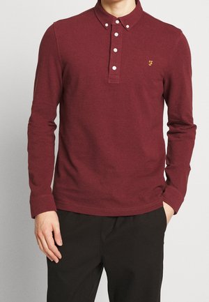 Homme portant un polo à manches longues bordeaux avec des boutons blancs et un pantalon noir, main dans la poche, debout devant un fond clair.
