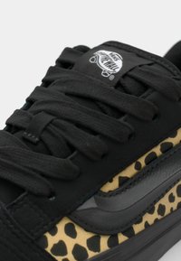 Svart sneaker med teksturert overdel, snøring og en leopardmønstret panel i beige med svarte prikker, supplert av en grå side stripe.