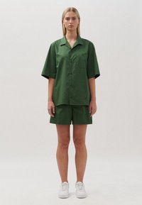 Completo composto da camicia verde a maniche corte e pantaloni corte in tessuto liscia. La camicia presenta un colletto e una tasca sul petto. Indossato con sneaker bianche.