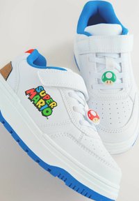 Weiße Sneaker mit einer blauen Gummisohle, Klettverschluss und Super Mario Grafiken. Zwei pilzförmige Akzente in Rot und Grün an den Schnürsenkeln.