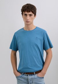 Ung mand iført en blå crew neck t-shirt og lyseblå jeans med et vævet brunt bælte, står med hænderne i lommerne mod en ensfarvet baggrund.