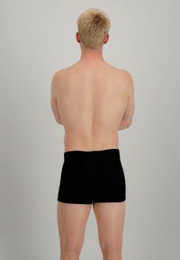 Shorts de bain noirs en tissu extensible, design ajusté avec une texture lisse, portés par une personne de dos, montrant un dos nu.
