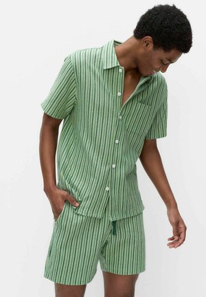 Jeune homme portant une chemise verte à manches courtes boutonnée et un short assorti avec des rayures verticales foncées, debout devant un fond blanc.