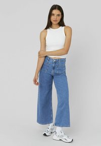 Top sans manches blanc associé à un jeans en denim bleu à taille haute et jambes larges. Le modèle porte des baskets blanches. Design simple avec deux poches arrière.