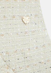 Witte textuurstof met zilveren en gouden draadaccenten, voorzien van een gouden hartvormig logo-label met 'Guess' gegraveerd.