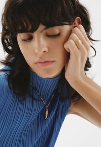 Collar de péndulo en forma de pez dorado sobre una cadena trenzada multicolor, combinado con una blusa azul de plisados que presenta un escote alto y una tela texturizada.