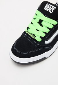 Černé semišové tenisky s jasně zelenými tkaničkami, bílým prošitím a logem Vans na jazyku. Obsahují bílý boční pruh a černou gumovou podrážku.