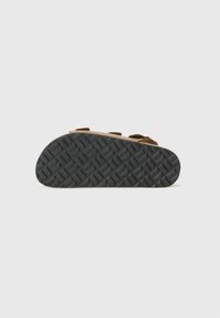 Sandal din suede maro, cu talpă plată, având trei curele ajustabile și o talpă din cauciuc texturat de culoare neagră. Partea inferioară are un model în diamant pentru aderență.
