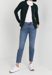 Woman wearing dark green cardigan over white turtleneck, blue jeans, and white sneakers standing against a plain background.