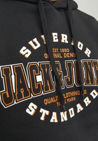 Černá mikina s kapucí obsahující velký bílý a oranžový grafický nápis "JACK & JONES" s texturovaným písmem, nastavitelné šňůrky a jemný povrch látky.