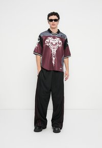 Μακρύ μπλουζάκι μπορντό και μαύρο με περίπλοκο λευκό γραφικό σχέδιο, συνδυασμένο με φαρδιά μαύρη παντελόνα με κόκκινες λεπτομέρειες και μαύρα μποτάκια.