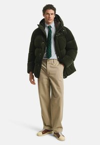 Dunkelgrüne Steppjacke mit Cord-Textur, kombiniert mit einem gestreiften Hemd und einer grünen Krawatte. Beige Hose mit braunen und creamfarbenen Sneakers.