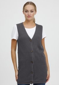 Grijze gebreide vest met een v-hals en knopen aan de voorkant. Heeft een gestructureerd, geweven patroon en wordt gedragen over een wit t-shirt.