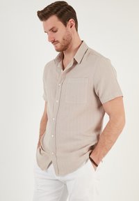 Camisa de manga corta de lino color beige claro, con un único bolsillo en el pecho y un corte relajado, combinada con pantalones blancos.