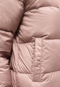 Κοντινό πλάνο ενός ροζ παλτού με ραφές, με μια τσέπη με καπάκι που ασφαλίζεται στο πλάι με ένα μόνο κουμπί πατιλέτα.