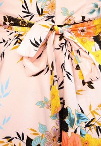 Kukallinen kangas, jossa on keltaisia, oransseja, sinisiä ja mustia kukkia, solmittuna nauhaksi vaaleanpunaista taustaa vasten.