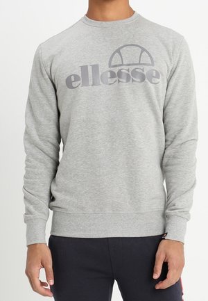 Homme portant un sweat-shirt gris clair avec un logo ellesse gris foncé sur la poitrine et un pantalon foncé.