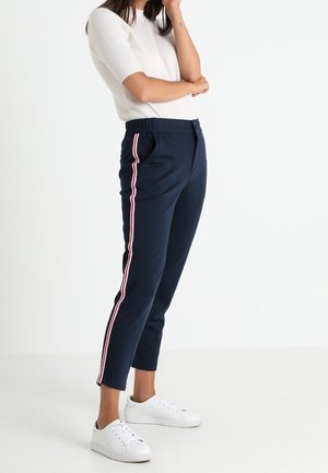 Kvinde iført navyblå bukser med røde og hvide side-striber, hvid kortærmet top og hvide sneakers, stående foran en hvid baggrund.
