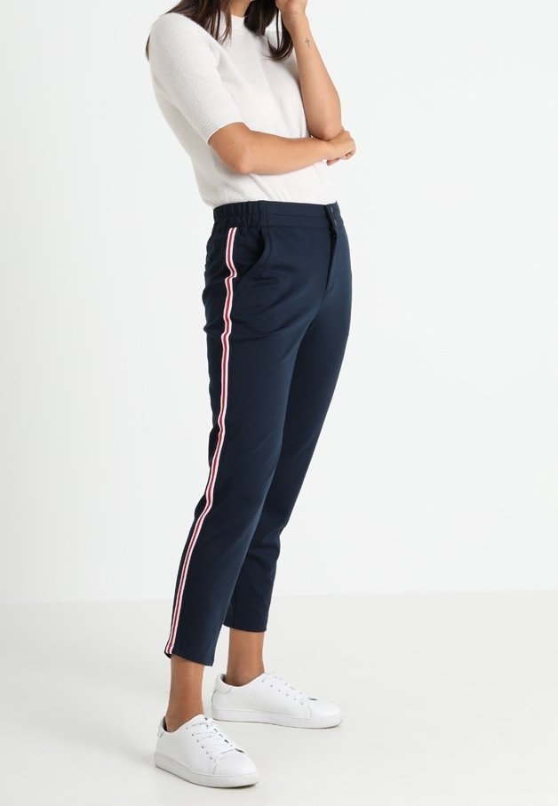 Femme portant un pantalon bleu marine avec des bandes latérales rouge et blanche, un haut blanc à manches courtes et des baskets blanches, se tenant contre un fond blanc.