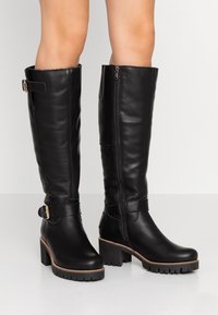 Botas de cuero negro hasta la rodilla con un tacón ancho, cremallera lateral y características de correa ajustable. Textura suave con suelas de goma.