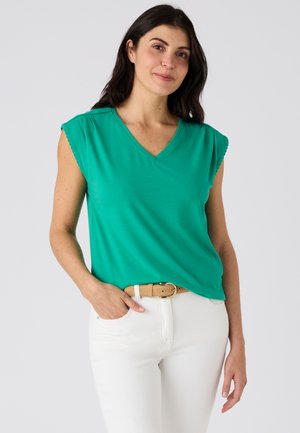 Vrouw met donker haar die een groen mouwloos V-hals topje, een witte broek en een beige riem draagt, staand met één hand in de zak.