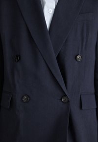 Blazer doppiopetto blu navy realizzato in tessuto testurizzato, dotato di due bottoni neri e una tasca sul lato sinistro. Camicia azzurro chiaro sotto.