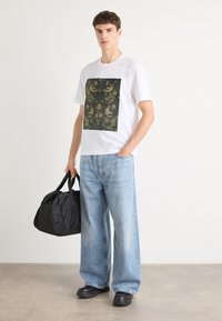 Weiße Baumwoll-T-Shirts mit einem schwarzen und goldfarbenen grafischen Muster; lockere, blaue Jeans; schwarze Handtasche; schwarze Schuhe.