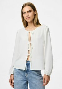 Weißes Bluse mit langen Ärmeln, Rundhalsausschnitt und vorderer Schleifenverschluss. Texturiertes Material und leichter Puff an den Schultern. Kombiniert mit blauen Jeans.