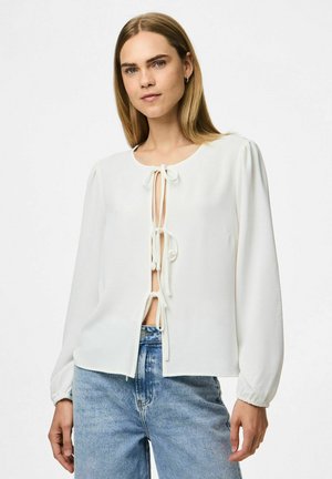 Blouse blanche à manches longues, col rond et fermeture par lien à l'avant. Tissu texturé et léger plissé aux épaules. Associée avec un jean bleu.