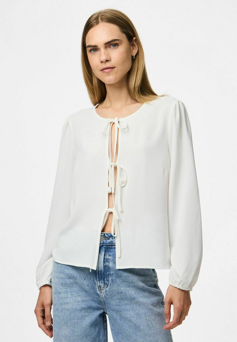 Hvid bluse med lange ærmer, rund hals og frontbinding. Tekstileret stof og let puff ved skuldrene. Kombineret med blå jeans.
