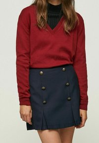 Pull rouge à col en V avec manches longues, associé à une jupe plissée bleu marine ornée de boutons dorés sur le devant et d'une finition texturée.