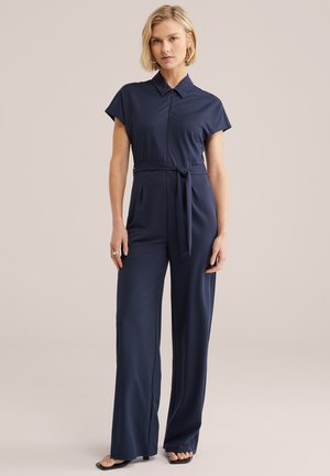 Lyshåret kvinde iført en marineblå korteærmet jumpsuit med krave og bælte i taljen, stående foran en ensfarvet beige baggrund.