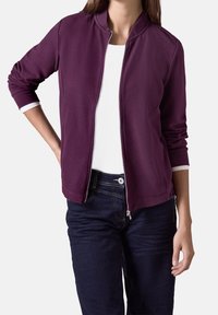 Veste zippée bordeaux avec col montant. Manches longues avec poignets blancs et texture lisse. Associée à un jean bleu foncé.