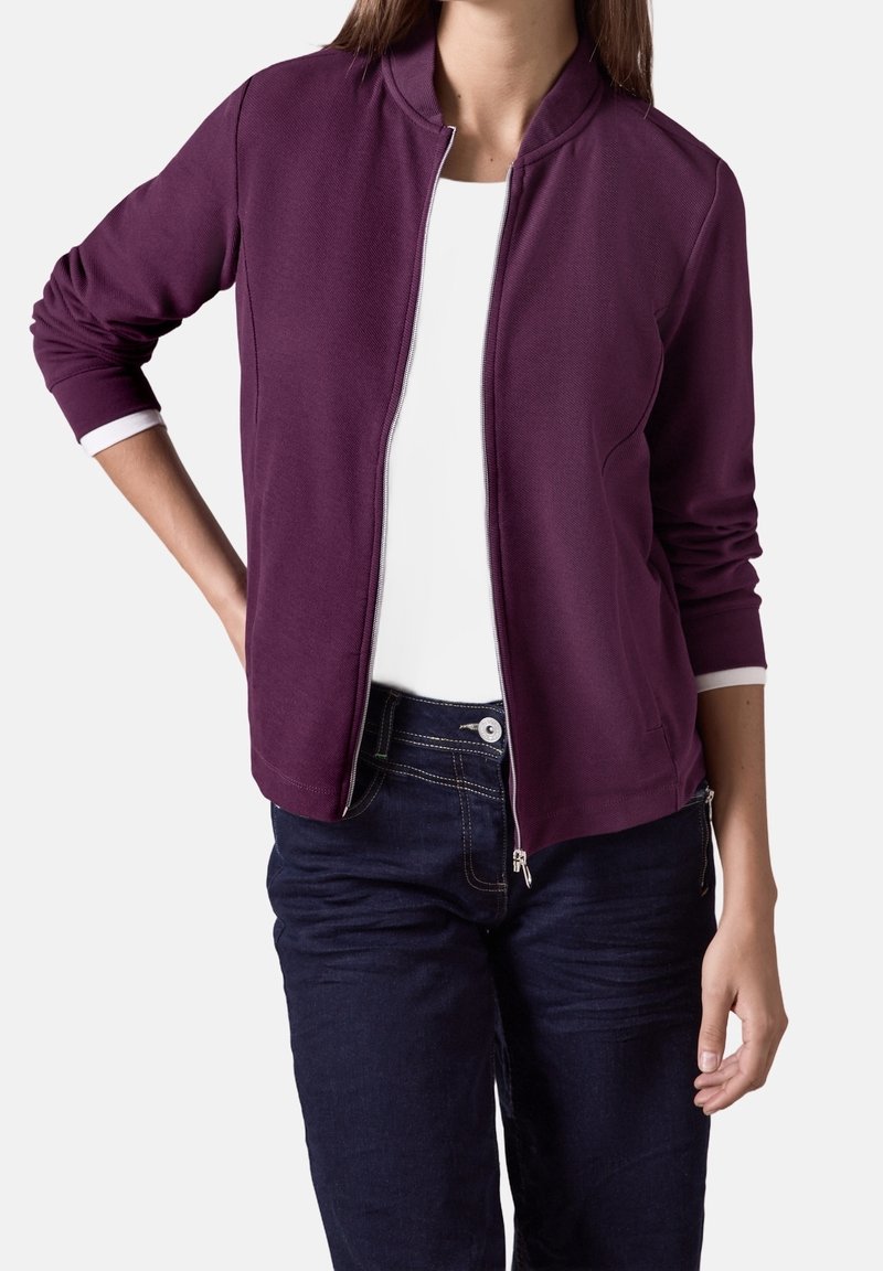 Veste zippée bordeaux avec col montant. Manches longues avec poignets blancs et texture lisse. Associée à un jean bleu foncé.