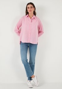 Light pink button-up shirt with long sleeves, relaxed fit, and tailored collar. Paired with light blue straight-leg jeans and white sneakers.