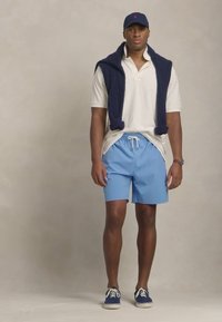 Model masculin îmbrăcat cu un tricou polo alb, pantaloni scurți albaștri deschiși și adidași bleumarin. Un pulover bleumarin din tricot cu cabluri este aruncat pe umeri.