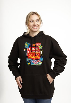 Smilende kvinde iført sort hættetrøje med farverig "Live Long and Prosper" Star Trek-grafik, hænderne i lommerne, stående mod hvid baggrund.