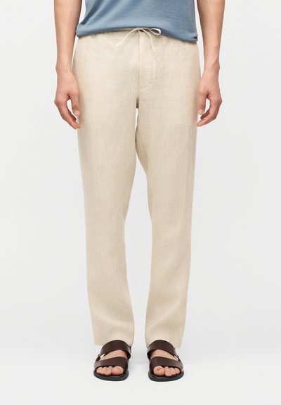 Homme portant un pantalon en lin beige avec une taille à cordon de serrage, une chemise bleue et des sandales ouvertes marron foncé, debout devant un fond uni.