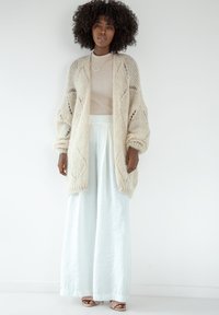 Kardigan pleciony w jasnym beżu, z otwartym przodem, w fakturowany wzór, w połączeniu z jasnoniebieskimi spodniami o szerokich nogawkach i dopasowaną beżową bluzką.