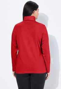 Červený rolák s dlouhým rukávem a jemnou texturou, s přiléhavým střihem a jemnými šicími detaily, doplněný černými kalhotami.