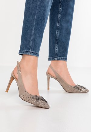 Füße, die beige Slingback-Pumps mit dekorativen Nieten tragen, kombiniert mit blauen Jeans vor einem schlichten weißen Hintergrund.