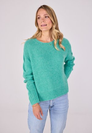 Türkiser Pullover aus weichem Material mit Rundhalsausschnitt, gerippten Bündchen und Saum, kombiniert mit hellblauen Jeans.