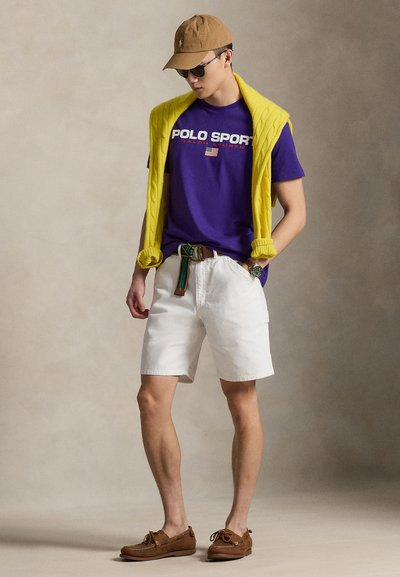 T-shirt en coton violet avec le logo "POLO SPORT", pull jaune en maille torsadée posé sur les épaules, short blanc, chaussures bateau marron et casquette kaki.