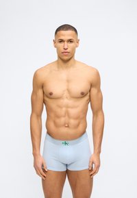 Νεαρός γυμνασμένος άνδρας που στέκεται με το πρόσωπο προς τα εμπρός, φοράει ανοιχτό μπλε ανδρικά εσώρουχα Calvin Klein boxer briefs, μπροστά σε απλό λευκό φόντο.