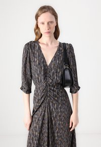 Schwarz-goldenes gemustertes Kleid mit tiefem V-Ausschnitt, gerafften Details und dreiviertellangen Puffärmeln. Das Model hält eine kleine schwarze Handtasche.