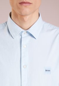 Světlounce modrá košile na knoflíky z bavlny. Má špičatý límec, knoflíky vpředu a malou čtvercovou nášivku s logem na levé straně hrudi.